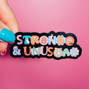 May include: A black sticker with the words "STRANGE & UNUSUAL" in colorful bubble letters. The design includes ghosts, skulls, and bats. The sticker is held against a pink background.