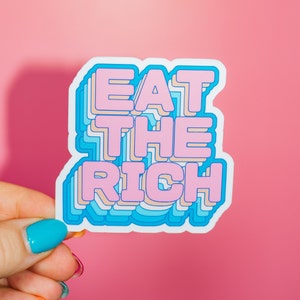 May include: A sticker with the phrase "Eat the Rich" in pink letters with a blue outline. The letters are layered and have a 3D effect.