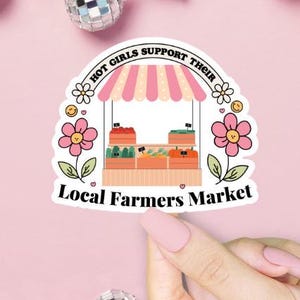 Pu&ograve; includere: Un adesivo rosa e bianco con un'illustrazione di un chiosco di un mercato contadino con il testo "Hot Girls Support Their Local Farmers Market".