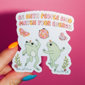 May include: White sticker with two green frogs dancing with a disco ball and butterflies. The sticker has the text "Be with people who match your energy."