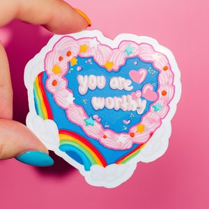 Pode incluir: Um adesivo em forma de cora&ccedil;&atilde;o com um design azul e rosa. O adesivo apresenta um arco-&iacute;ris, nuvens e o texto "You are worthy" em uma fonte rosa e branca.