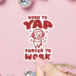 May include: Red sticker with a white outline featuring a cartoon cat wearing headphones and singing. The text on the sticker reads "Born to Yap Forced to Work".
