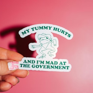 May include: White sticker with green text that reads "My tummy hurts and I'm mad at the government". A cartoon frog wearing a cowboy hat is in the center of the sticker.