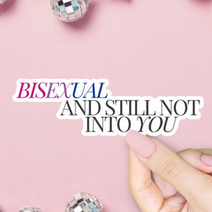 May include: A white sticker with the text "BISEXUAL" in pink and blue, and "AND STILL NOT INTO YOU" in black. The sticker is held by a hand with pink nails against a pink background, with disco balls.