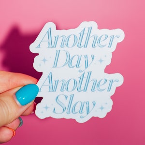 May include: A white sticker with the text "Another Day Another Slay" in light blue script. The sticker has a decorative border and star accents. The background is a solid pink.