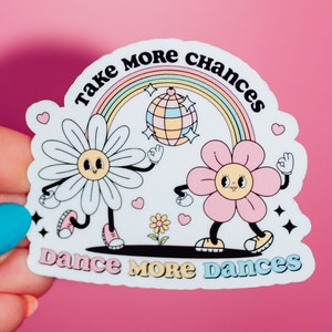 May include: White sticker with a rainbow, a disco ball, and two cartoon flowers with the text "Take More Chances Dance More Dances".