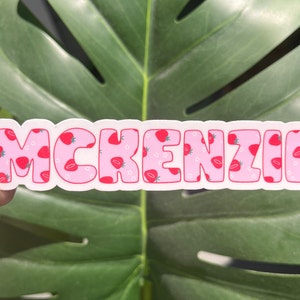 May include: A sticker with the name "MCKENZIE" in bubble letters. The letters are pink with a strawberry pattern and white outlines. The background is a green leaf.