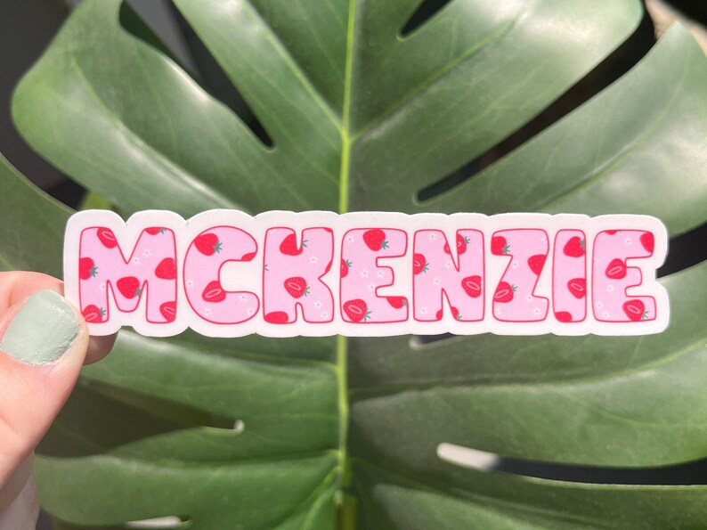 May include: A sticker with the name "MCKENZIE" in bubble letters. The letters are pink with a strawberry pattern and white outlines. The background is a green leaf.