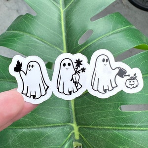 May include: Three black and white ghost stickers on a green leaf. The ghosts are in different poses, one holding a flower, one holding a watering can, and one holding a butterfly.