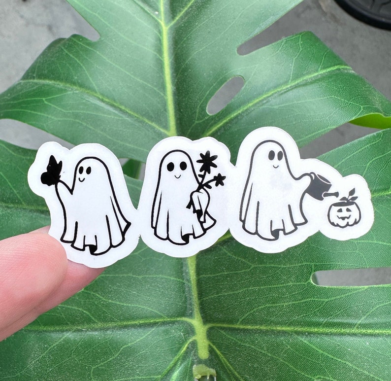 May include: Three black and white ghost stickers on a green leaf. The ghosts are in different poses, one holding a flower, one holding a watering can, and one holding a butterfly.