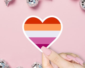 May include: A heart-shaped sticker with the lesbian pride flag design, featuring orange, white, pink, and magenta stripes. The sticker is held against a pink background with several small disco ball ornaments.
