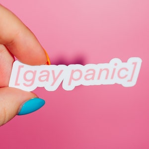 May include: A white sticker with pink text that reads "[gay panic]" being held by a hand with blue and orange nail polish.