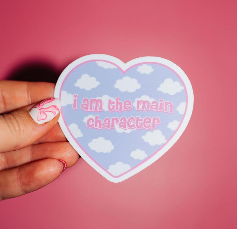 May include: A heart-shaped sticker with a blue sky and white clouds background. The sticker has pink text that reads "i am the main character".