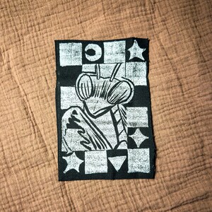 May include: A black and white patch featuring a stylized insect design. The insect has large, detailed eyes and is set against a checkered background with star and geometric shapes. The patch is rectangular.