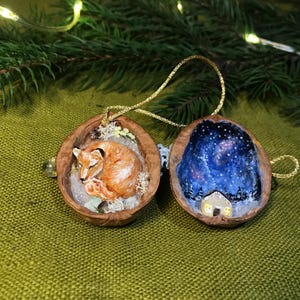 May include: Two walnut shell halves used as hanging ornaments with gold strings. One shell contains a painted orange fox curled up on white material. The other shell features a painted night sky with stars and a small house with glowing windows.