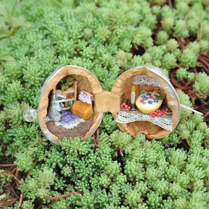 May include: A miniature scene created inside two halves of a walnut shell. One half shows a badger figurine sitting in a room with a bookshelf and a bed. The other half shows a miniature table with a candle and a blueberry pie.