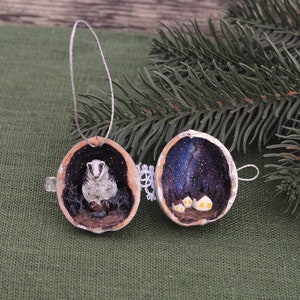 May include: A pair of hand-painted walnut shell ornaments. One ornament features a badger inside a hollowed-out walnut shell with a starry night sky painted on the inside. The other ornament features a starry night sky with a small village painted on the inside.