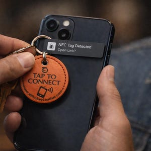 以下が含まれることがあります： 「TAP TO CONNECT」の文字とスマートフォンのアイコンが入ったオレンジ色の革製キーホルダーが、黒いスマートフォンの近くに持たれています。携帯電話には、NFCタグが検出されたことを示す通知が表示されています。