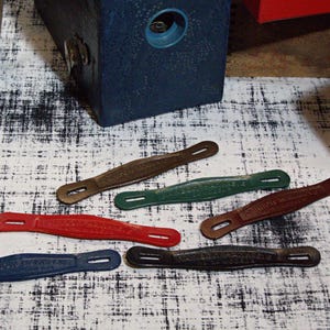 Könnte beinhalten: Eine Sammlung von Vintage-Ledergriffen in verschiedenen Farben, darunter Rot, Grün, Braun und Blau. Jeder Griff hat eine unverwechselbare Form mit einer zentralen Öffnung und ist wahrscheinlich für einen Koffer oder eine Reisetasche bestimmt. Die Griffe sind auf einer gemusterten Oberfläche angeordnet.