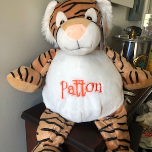 May include: A plush tiger toy with orange and black stripes. The toy has a white belly with the name "Patton" embroidered in orange thread.
