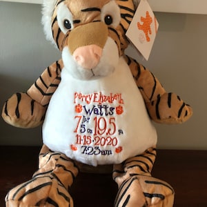 May include: A plush tiger stuffed animal with an embroidered white shirt. The shirt says "Perry Elizabeth Watts 7 lbs 5 oz 19.5 in 11.15.2020 7:23 am" in purple and orange thread.