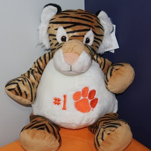 May include: A plush tiger stuffed animal wearing a white shirt with an orange paw print and the number "#1" embroidered on it. The tiger is sitting on an orange surface.