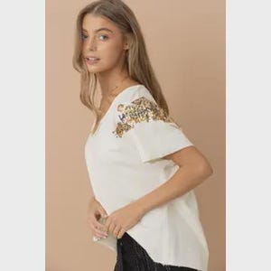 May include: Cream-colored V-neck t-shirt with short sleeves. The shoulder area is embellished with a floral sequin design in gold, brown, and green. The model is wearing the shirt.