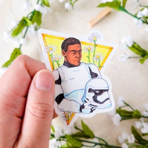 May include: A sticker featuring a cartoon illustration of a Star Wars character wearing a white helmet and holding a white helmet. The character has brown skin and is surrounded by white daisies.