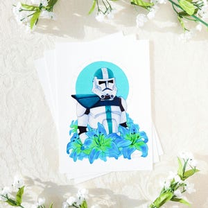 May include: A white and blue Star Wars clone trooper with a teal helmet stands in a field of blue flowers. The trooper is surrounded by blue lilies and has a teal stripe down the center of its armor.