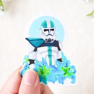 May include: A sticker featuring a white and green Star Wars clone trooper with a blue and green halo and surrounded by blue and green lilies.