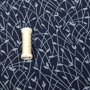 May include: A spool of off-white thread sits on a navy blue fabric with a light blue leaf and stem pattern. The fabric has a textured appearance, and the thread spool is centered in the image.