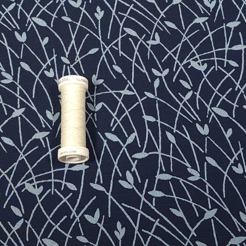 May include: A spool of off-white thread sits on a navy blue fabric with a light blue leaf and stem pattern. The fabric has a textured appearance, and the thread spool is centered in the image.