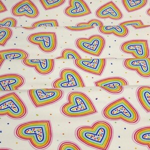 K&ouml;nnte beinhalten: Wei&szlig;er Stoff mit einem sich wiederholenden Muster aus regenbogenfarbenen Herzen. Jedes Herz ist rosa und blau umrandet und hat ein gepunktetes Design in der Mitte. Kleine, bunte Punkte sind &uuml;ber den Hintergrund verstreut.