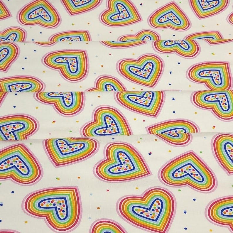 K&ouml;nnte beinhalten: Wei&szlig;er Stoff mit einem sich wiederholenden Muster aus regenbogenfarbenen Herzen. Jedes Herz ist rosa und blau umrandet und hat ein gepunktetes Design in der Mitte. Kleine, bunte Punkte sind &uuml;ber den Hintergrund verstreut.
