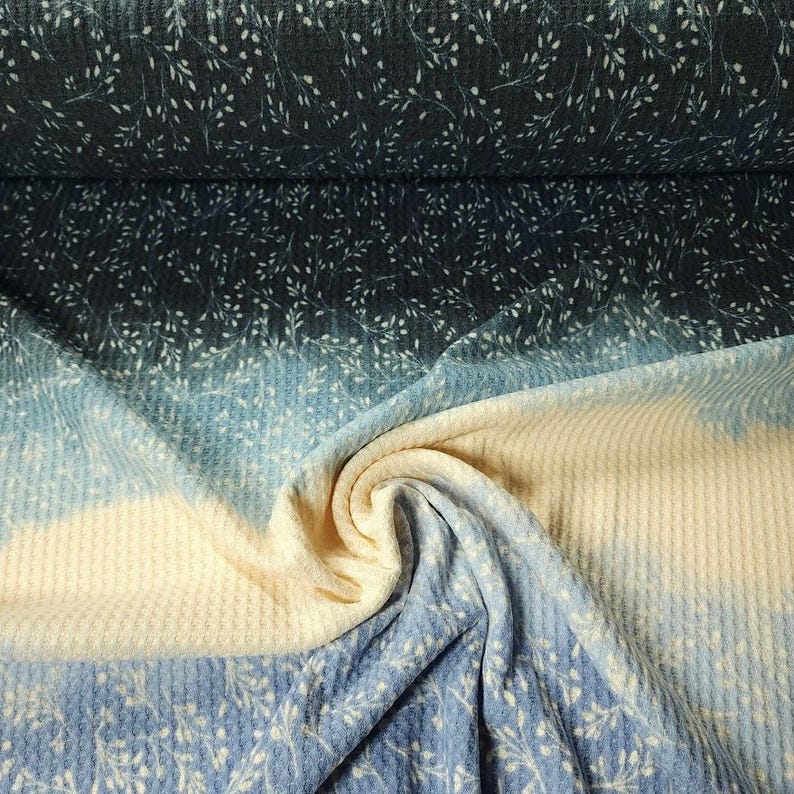 May include: A roll of fabric with a gradient effect, shifting from dark blue to light blue and cream. The material has a waffle weave and a subtle white floral design. Ideal for sewing and craft projects, the fabric is versatile.