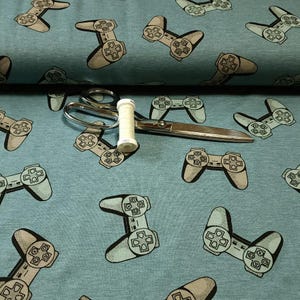 May include: Light blue fabric with a repeating pattern of gray and black video game controllers. A pair of silver scissors and a spool of white thread are on the fabric.
