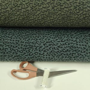 May include: Two rolls of patterned fabric in shades of green and teal, with a dark, irregular design. A pair of peach-colored scissors and a spool of white thread are in the foreground, suggesting sewing or crafting.