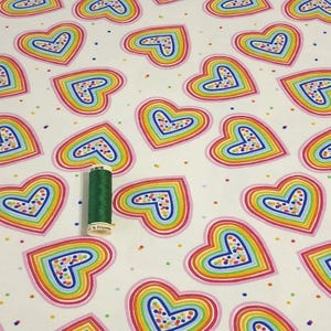 K&ouml;nnte beinhalten: Wei&szlig;er Stoff mit einem sich wiederholenden Muster aus regenbogenfarbenen Herzen. Jedes Herz hat einen rosa Umriss mit einem blauen Umriss im Inneren. Eine gr&uuml;ne Garnrolle liegt auf dem Stoff. Die Herzen sind mit bunten Punkten gef&uuml;llt.
