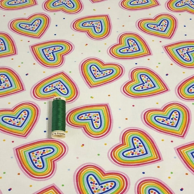 K&ouml;nnte beinhalten: Wei&szlig;er Stoff mit einem sich wiederholenden Muster aus regenbogenfarbenen Herzen. Jedes Herz hat einen rosa Umriss mit einem blauen Umriss im Inneren. Eine gr&uuml;ne Garnrolle liegt auf dem Stoff. Die Herzen sind mit bunten Punkten gef&uuml;llt.