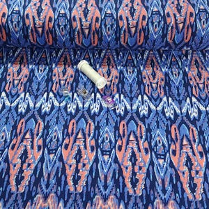 May include: A bolt of patterned fabric in shades of blue and coral. The fabric features a repeating ikat design. A white spool of thread and three colorful sewing clips are on the fabric.