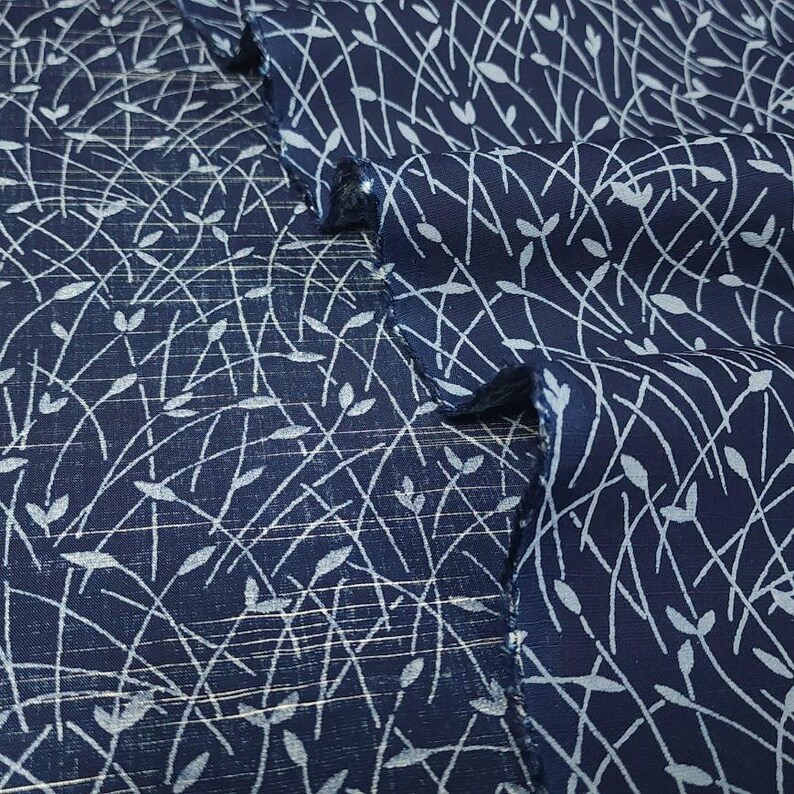 May include: Close-up of navy blue fabric with a white, delicate, linear pattern. The design features thin, curved lines and small leaf-like shapes, creating a textured, organic aesthetic. The fabric is folded, showing the pattern's repeating nature.