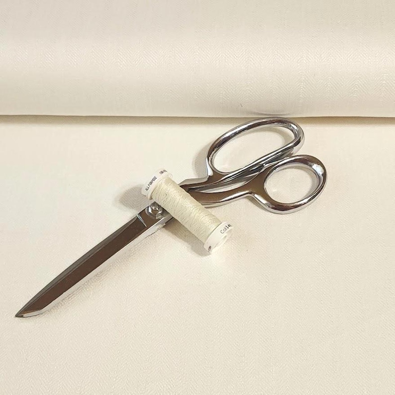 以下が含まれることがあります： 銀色の裁ちばさみと、クリーム色の糸巻き。 はさみは開いており、糸巻きが刃の間に置かれています。 糸巻きには白い文字が書かれています。 背景は明るいクリーム色です。