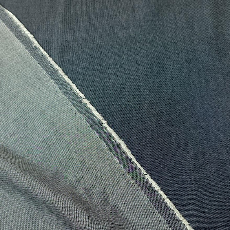 Puede incluir: Primer plano de dos secciones de tela vaquera. Una secci&oacute;n est&aacute; doblada, revelando un tono azul m&aacute;s claro en la parte inferior. Los bordes est&aacute;n deshilachados y la tela tiene un aspecto texturizado. El color general es un &iacute;ndigo profundo.