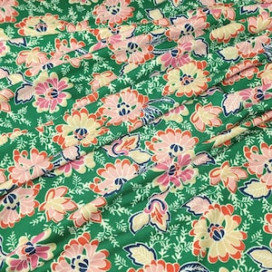 May include: A green fabric featuring a repeating floral pattern with large stylized flowers in shades of light pink, cream, and orange, accented by dark blue centers and small white leafy vine details throughout the textile surface.