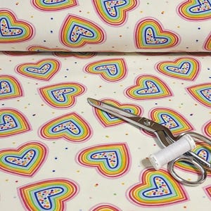 K&ouml;nnte beinhalten: Wei&szlig;er Stoff mit einem sich wiederholenden Muster aus regenbogenfarbenen Herzen. Die Herzen sind rosa umrandet und mit bunten Punkten gef&uuml;llt. Eine silberne Schere und eine Spule wei&szlig;en Fadens liegen auf dem Stoff.