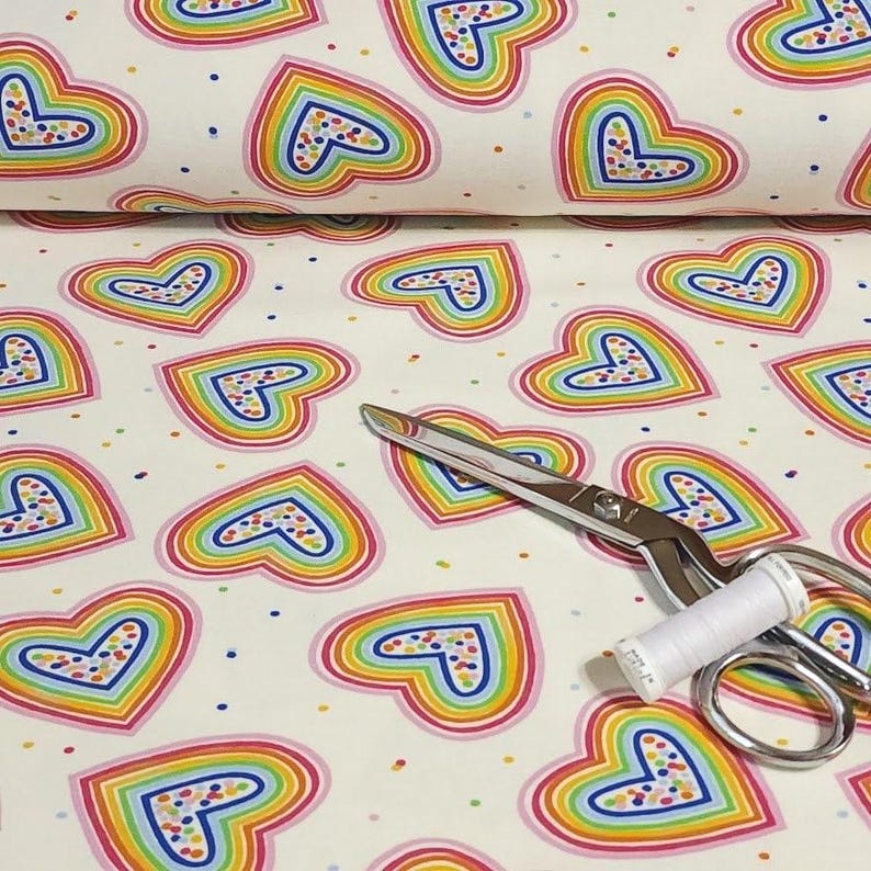 K&ouml;nnte beinhalten: Wei&szlig;er Stoff mit einem sich wiederholenden Muster aus regenbogenfarbenen Herzen. Die Herzen sind rosa umrandet und mit bunten Punkten gef&uuml;llt. Eine silberne Schere und eine Spule wei&szlig;en Fadens liegen auf dem Stoff.