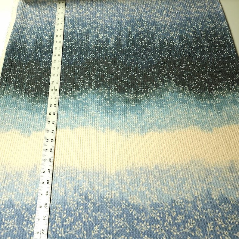 May include: Blue and white fabric with a gradient design. The fabric has a ribbed texture and a pattern of small white floral sprigs. A ruler is placed vertically along the fabric, showing measurements in inches.