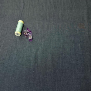 Puede incluir: Una bobina de hilo verde claro y una pinza de costura morada descansan sobre una tela gris oscuro. La bobina de hilo tiene una etiqueta amarilla. La pinza de costura es de pl&aacute;stico transparente y tiene un mecanismo de resorte. La tela tiene una textura sutil.