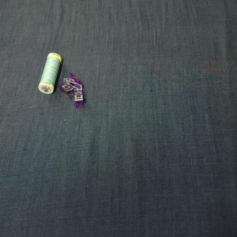 Puede incluir: Una bobina de hilo verde claro y una pinza de costura morada descansan sobre una tela gris oscuro. La bobina de hilo tiene una etiqueta amarilla. La pinza de costura es de pl&aacute;stico transparente y tiene un mecanismo de resorte. La tela tiene una textura sutil.