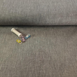 May include: A roll of gray linen fabric with a textured surface. A white thread spool and four colorful sewing clips are scattered on the fabric. The clips are yellow, purple, and blue.